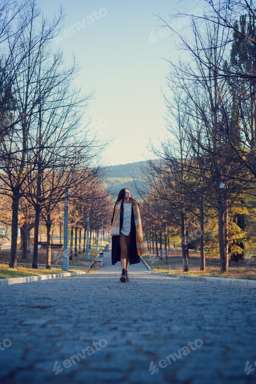 Preview: Woman in an overcoat and scarf walks through an autumn park at dusk