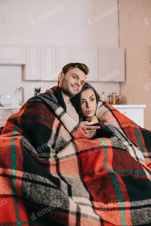 Preview: emotional young couple relaxing on couch and watching tv together while covering with plaid