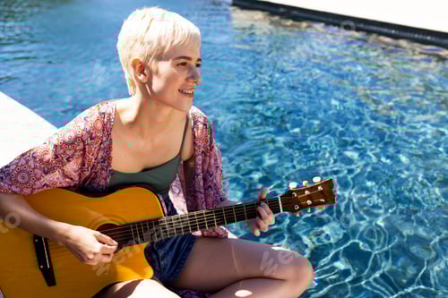 Preview: Non-binary musician sitting poolside in sun, playing acoustic guitar near clear water, copy space