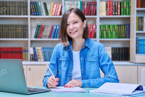 Preview: Young female university student looking at camera with laptop in library