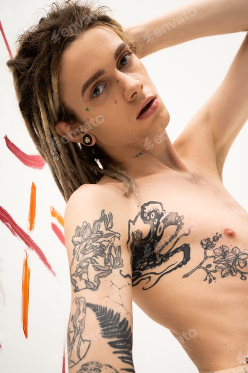 Preview: young tattooed nonbinary model with dreadlocks looking at camera near colorful paint strokes on