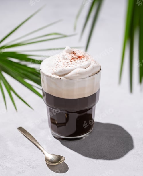 Preview: Iced coffee in a glass with foam on a light background with palm leaves, spoon and morning shadows.