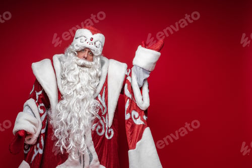 Preview: Santa Claus elderly male in festive xmas costume point by hand to copy space on red background