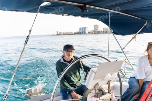 Preview: Senior man steering sailboat with woman on board near coastline