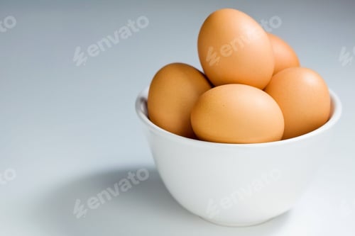 Preview: White bowl containing brown eggs placed atop a table, against the pale backdrop