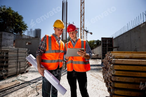 Preview: Civil architect and construction manager dressed in orange work vests and helmets discuss a building
