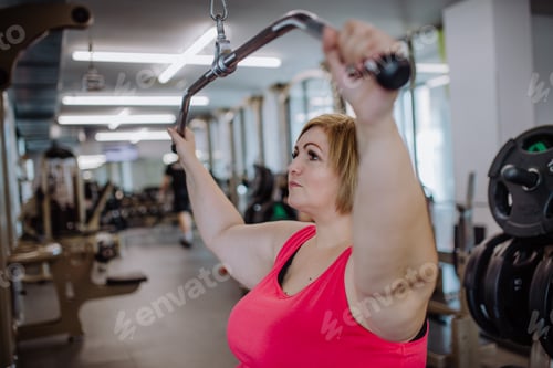 Preview: Mid adult plus size woman working out on a lat pulldown machine indoors in gym