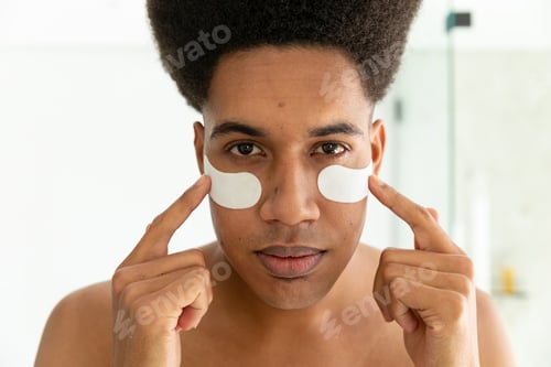 Preview: Biracial man looking in mirror and applying under eye masks in sunny bathroom