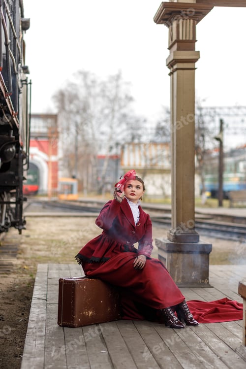 Preview: A beautiful girl in a burgundy suit is sitting on a suitcase near an old steam locomotive