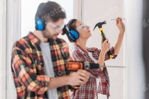 Preview: young couple in protective workwear hammering and drilling wall