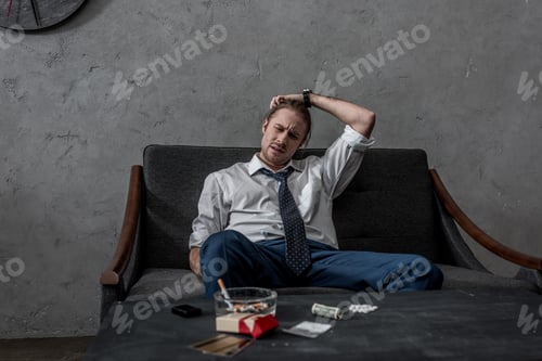 Preview: depressed businessman with drug addiction sitting on couch in front of table with drugs