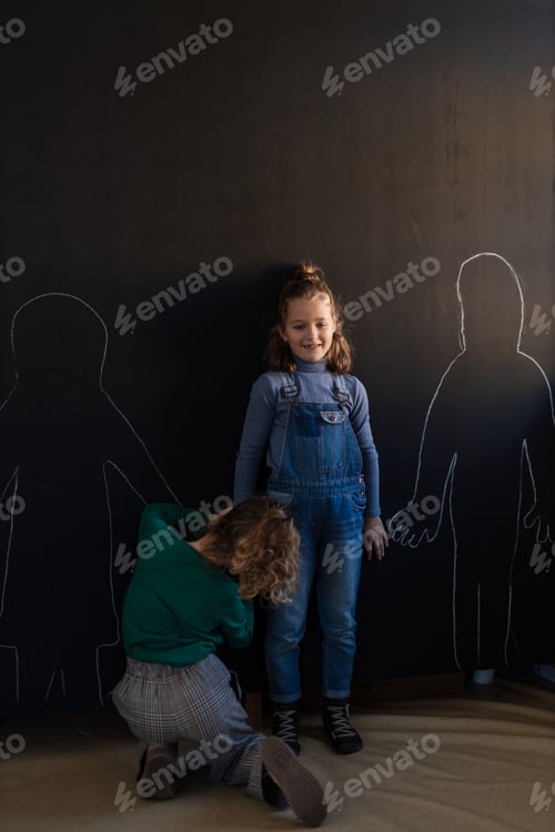 Preview: Rear view of little girl drawing her friends with chalks on blackboard wall indoors in playroom