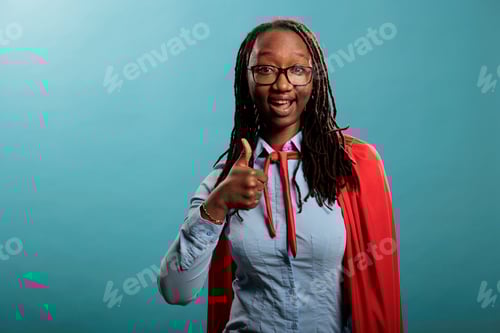 Preview: Brave and proud superhero woman wearing cloak while giving thumbs up gesture sign on blue background