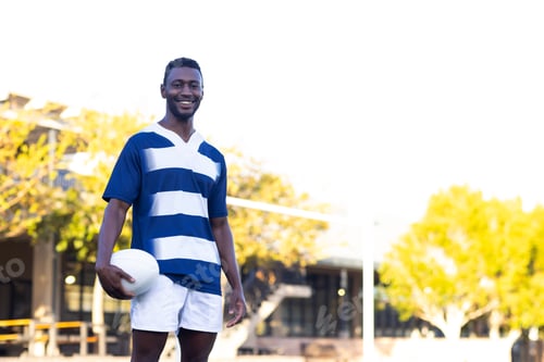 Preview: African American young male athlete holding a rugby ball, standing on field outdoors, copy space