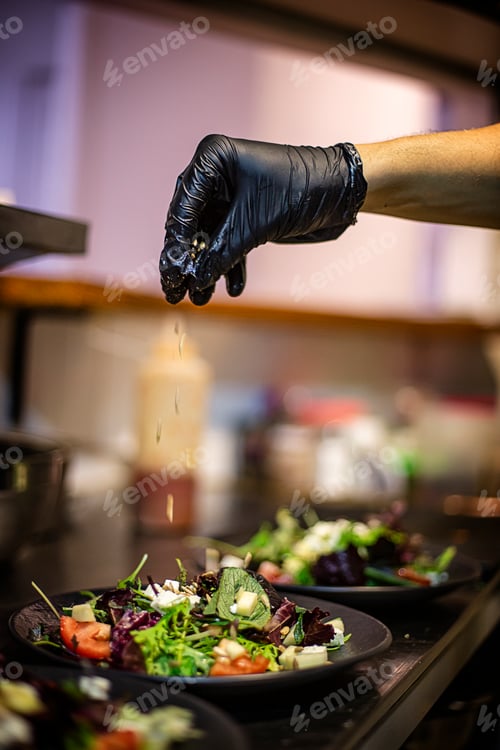 Preview: Cook's hand with black plastic glove dropping seeds on a salad in a restaurant kitchen.