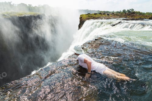Preview: Man in the water at the Devils Pool on the edge of Victoria Falls, looking over the waterfall edge.