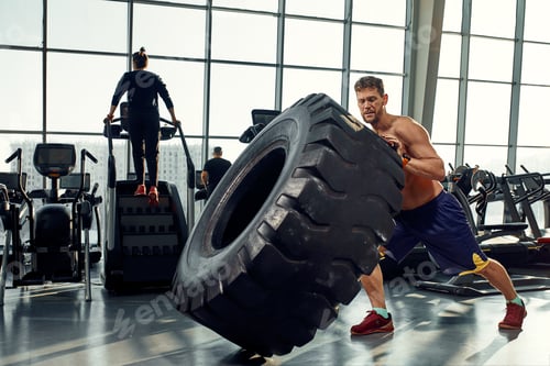 Preview: Strongman, bodybuilding, sport, fitness and people concept - young man doing tire flip at training