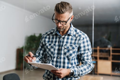 Preview: Serious white-haired man working with papers while standing in office
