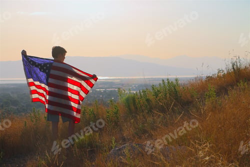 Preview: Young boy holding American flag behind standing on mountain peak overlooking valley at golden hour.