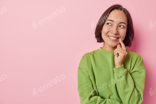 Preview: Horizontal shot of pretty dreamy Asian woman with short dark hair smiles happily keeps finger near c