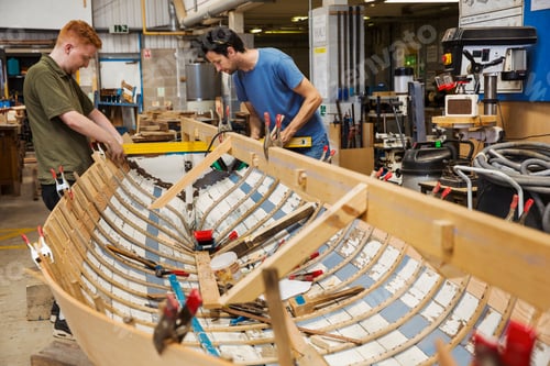 Preview: Two men in a boat-builder's workshop, working together on a wooden boat hull.