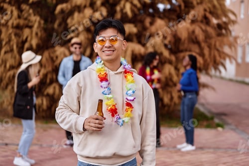Preview: Asian man with a beer bottle smiling while having a party with friends outdoors.