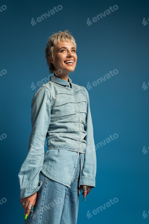 Preview: Charming young woman in stylish denim outfit smiles against a vibrant blue background