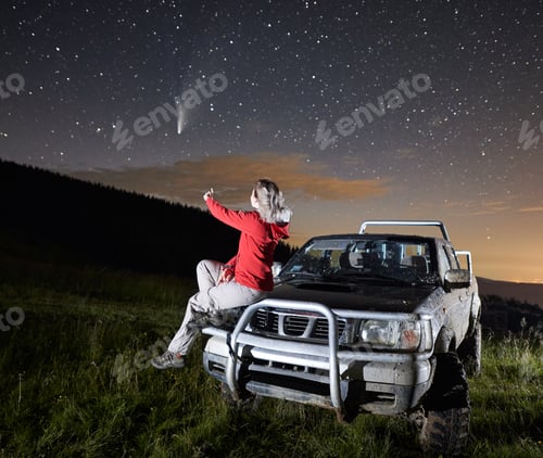Preview: Young woman in the mountains watching beautiful starry night and a comet, sitting on car hood