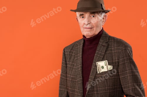 Preview: trendy senior man in hat standing with money in pocket isolated on orange