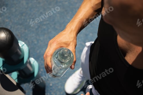 Preview: Glass bottle of refreshing water in athletes man hand on sports ground. Water balance for workout.