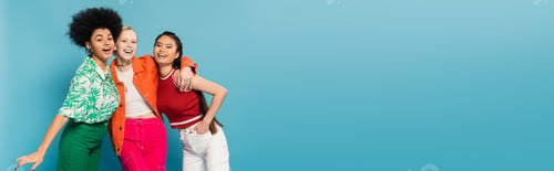 Preview: joyful multicultural women in trendy and colorful clothes embracing and smiling at camera isolated