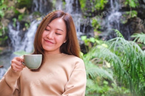 Preview: A young woman holding and drinking coffee while sitting in the garden with waterfall