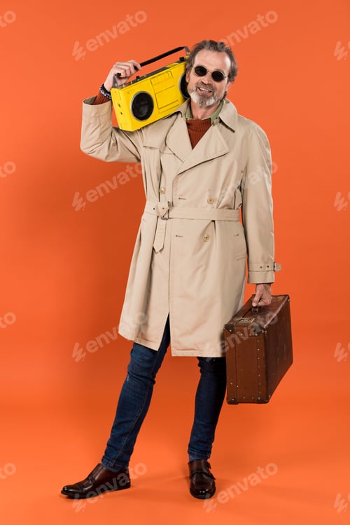 Preview: cheerful retired man holding yellow boombox and briefcase in hands on orange background