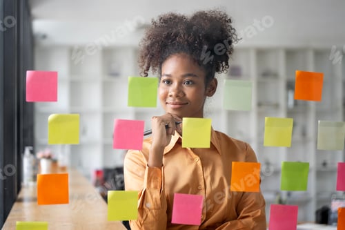 Preview: Young businesswoman using sticky notes in glass wall to writing strategy business plan to success