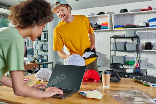 Preview: Smiling young designer holding snapbacks while talking to african american colleague using laptop