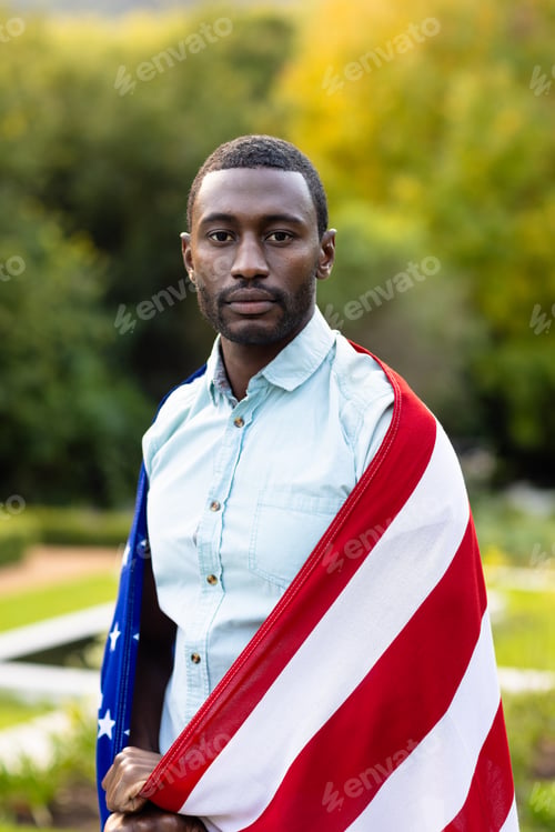 Preview: Portrait of serious african american man covered with flag of usa in garden