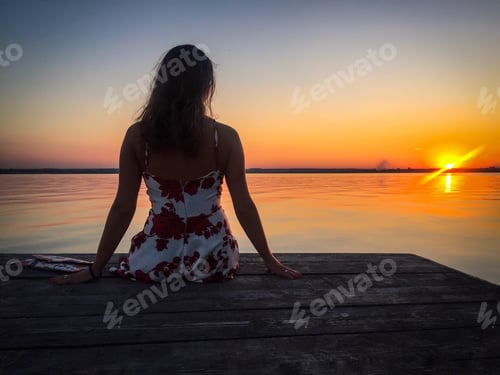 Preview: Back of woman wearing summer dress on wooden pontoon watching the sunset over the lake