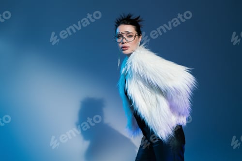 Preview: young woman in transparent eyeglasses and fluffy faux fur jacket looking at camera on blue