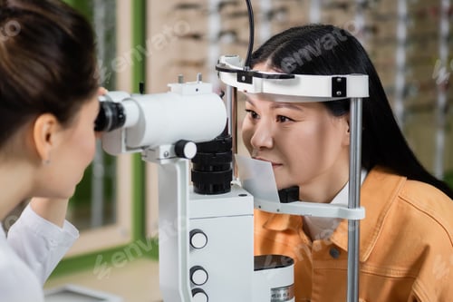 Preview: blurred optometrist testing eyesight of asian woman on vision screener in optics shop