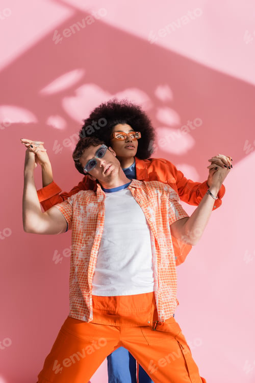 Preview: Stylish african american woman holding hands of brunette man in overall on pink background with