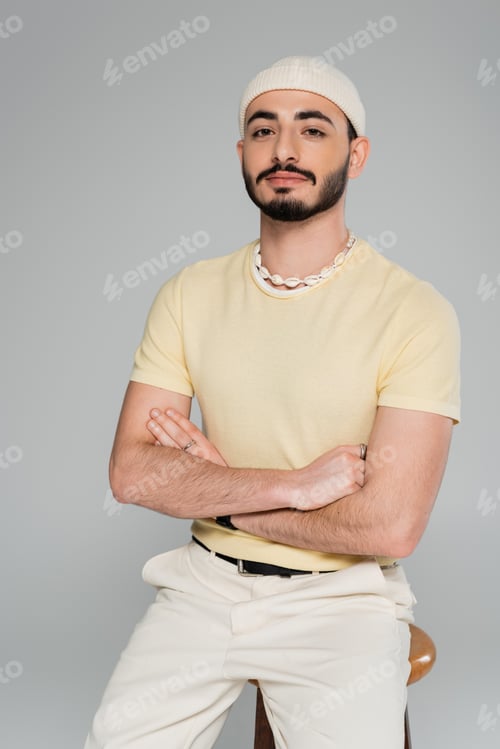 Preview: portrait of trendy gay man in hat crossing arms near chair isolated on grey
