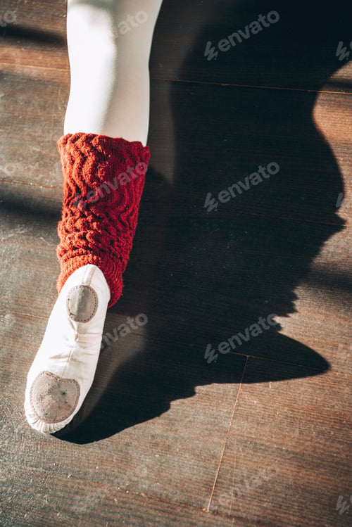 Preview: Crop shot from above of ballerina's leg wearing white pointe shoe on wooden floor.