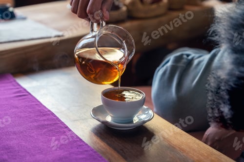 Preview: Hand pouring hot tea from jug into white teacup on the table