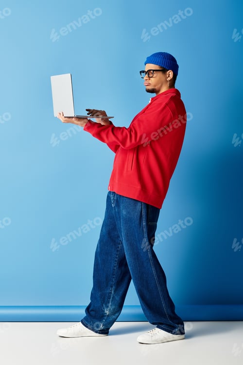 Preview: Trendy young man in a beanie works on a laptop against a vibrant blue backdrop