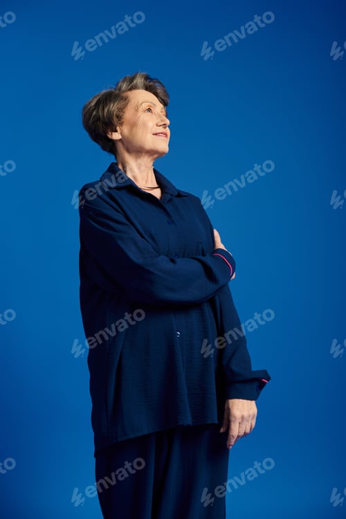 Preview: A confident woman in her 60s poses gracefully in trendy attire against a vibrant blue backdrop.