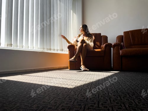Preview: Woman sitting on the sofa near the window and looking through the blinds
