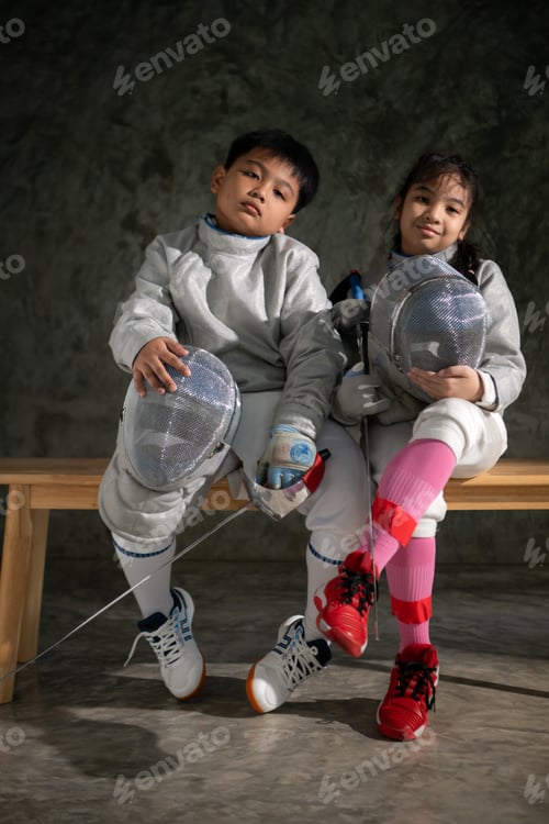 Preview: Two child in fencing gear sit on a bench, smiling at each other