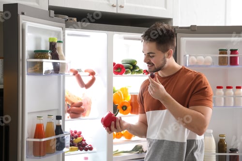 Preview: Young man with bell pepper and grape berry near open refrigerator indoors