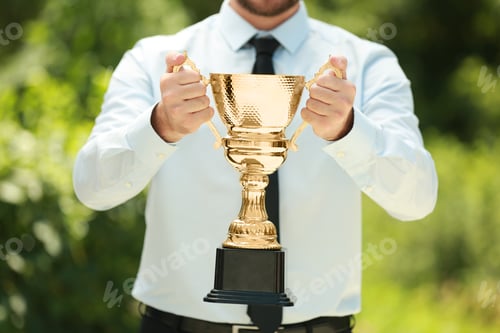 Preview: Young businessman holding gold trophy cup in green park, closeup