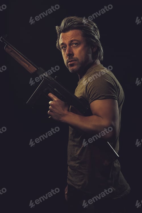 Preview: Rugged Man With Gun In Green T-Shirt Against Dark Background.